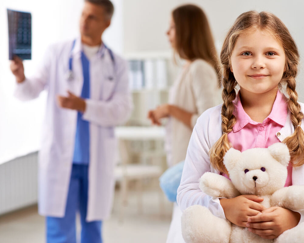 Happy girl with teddy bear looking at camera on background of doctor showing x-ray results to her mother